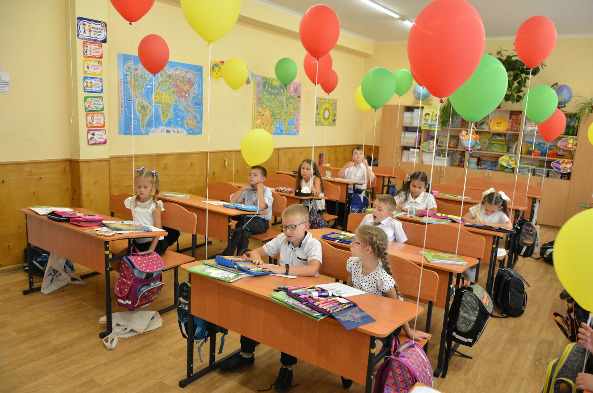 Children sitting at desks in a classroom decorated with balloons.